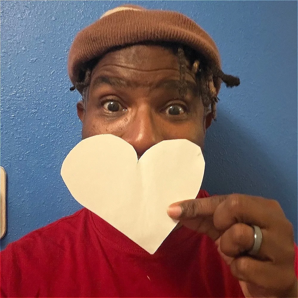 A man in a brown woolly hat and red top holds a white paper heart over his mouth against a blue wall